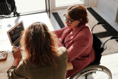 Person in a wheelchair working at a desk engaging in conversation with another person.