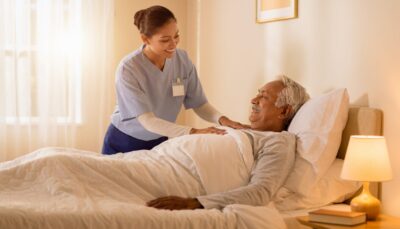 A warm, softly lit bedroom scene showing an elderly person resting comfortably in bed, covered by clean white bedding. A caring nurse or support worker is gently attending to them, adjusting the covers or checking on the patient. The bed has a subtle medical air mattress or pressure care overlay visible beneath the bedding. Natural light streams through a sheer-curtained window. The room feels calm, domestic, and dignified — not clinical or institutional. A small bedside lamp adds warmth. The overall mood is reassuring, compassionate, and peaceful. Editorial healthcare photography style, shallow depth of field, warm colour palette of cream, soft white and pale gold tones.